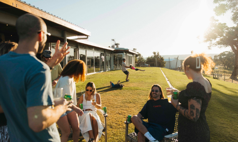A group of friends relaxing on a sunny day on a grassy lawn next to a modern house. Several people are sitting in lawn chairs holding green drinks, while one person stands up on the right in a black dress.