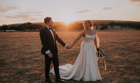 A happy couple who just got married hold hands at the sun sets in the background of a grassy, rustic field at Eagle Bay's wedding venue