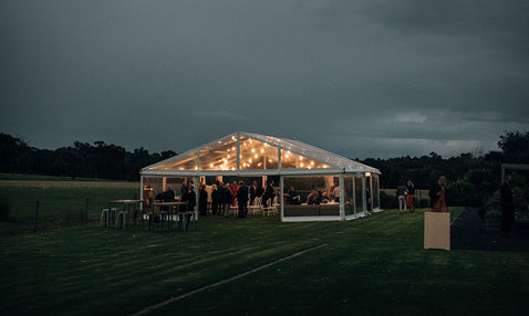 A lit-up wedding marquee sits at a distance with wedding guests inside at night time at Eagle Bay Brewing Co's wedding venue