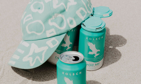 A mint green Kolsch Eagle Bay can with a flip-top lid sits in sand next to a matching mint green Kolsch cap and another can, all featuring a white eagle logo and pattern.