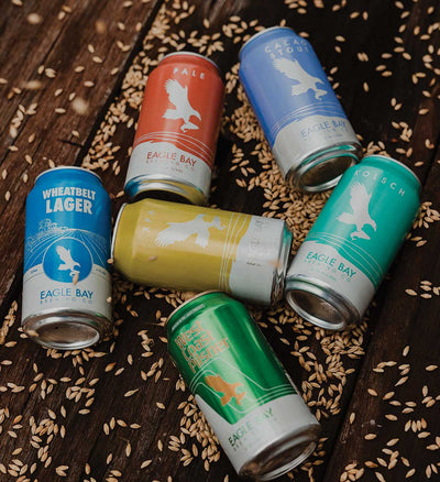 Collection of Eagle Bay Brewing Company beer cans on a wooden surface with grain.