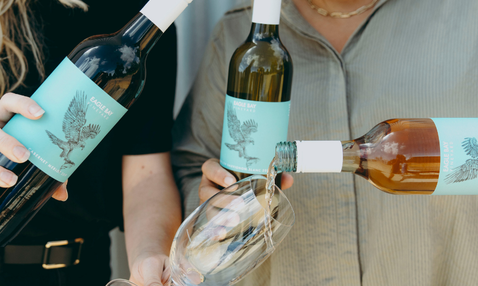 Three wine bottles with light blue labels are being held by people, with one bottle pouring wine into a glass. The scene is cropped closely and shows the bottles and pouring action in a real-life setting.